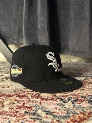 Svart White Sox keps från New Era - Säljer en svart keps från New Era med White Sox-logga broderad i vitt framtill och ett litet baseballemblem. På sidan finns ett broderat 'World Series 05'-märke. Klassisk 59Fifty-modell med rak skärm och broderad New Era-logga på sidan.