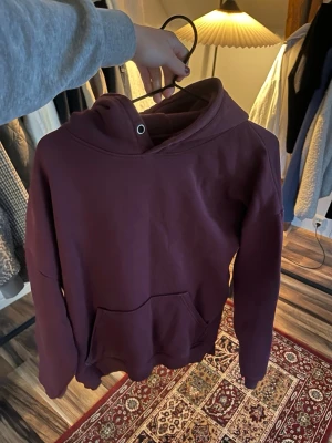 Vinröd hoodie med magficka - Säljer en vinröd hoodie med klassisk huva och stor magficka framtill. Hoodien har ribbade muddar vid ärmslut och nederkant. Perfekt för dig som gillar en enkel och stilren look. Materialet känns mjukt och skönt mot huden.