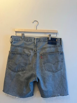 Levi's ljusblå jeansshorts med rå kant - Ljusblå jeansshorts från Levi's med klassisk femficksdesign och rå, avklippt kant nertill. Shortsen har normal passform och midja, samt tydliga Levi's-detaljer som den röda loggan på bakfickan. Perfekta för varma dagar och enkel att styla.
