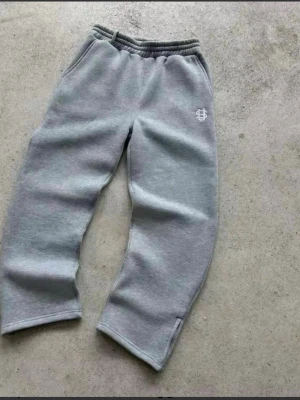 Suvene Grå mjukisbyxor - Säljer ett par grå mjukisbyxor från Suvene med broderad vit logga på vänster lår och vid benslutet. Byxorna har avslappnad passform, resår i midjan och praktiska sidofickor. Perfekta för chill dagar och streetwear-stil.