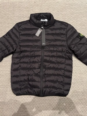 Svart pufferjacka från Ston Island - NY OANVÄND MED PRISTAGG KVAR! Snygg svart pufferjacka från Ston Island med klassisk patch på vänster ärm. Jackan har ståkrage, dragkedja framtill och quiltad design. Perfekt för kalla dagar och riktigt stilren look. Materialet är lätt och isolerande. OBS! Det står att den har storlek XL, men den är snarare en liten LARGE eller en stor MEDIUM.