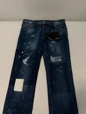 Blå distressed jeans från Dsquared2 - Säljer ett par blå jeans från Dsquared2 med coola slitningar, färgstänk och patchdetaljer. Jeansen har en relaxed fit och klassisk femficksdesign. Perfekta för dig som gillar streetwear och vill sticka ut med en unik look.             Tar jättegärna emot byten!!