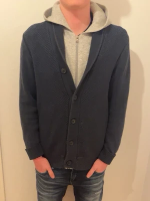 Mörkblå kofta med knappar - Stilren mörkblå kofta med grovstickad struktur och stora knappar framtill. Koftan har ribbade muddar vid ärmslut och nederkant samt en klassisk V-ringning. Perfekt att bära över en hoodie för en avslappnad look. Skriv för mer info