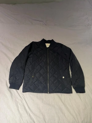 Holly & Whyte jacka (barn) - Snygg svart quiltad bomberjacka från Holly & Whyte med dragkedja framtill, ribbade muddar och två fickor med knapp. Jackan har ett klassiskt bomberkrage och är tillverkad i ett glansigt, lätt material som ger en stilren look. Passar perfekt som vår jacka, tunn och lätt. Den är i storlek 146.