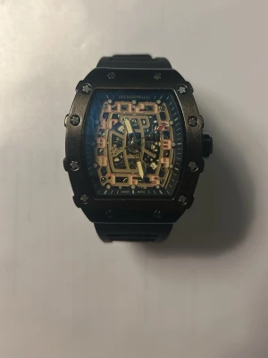 RM svart klocka - Snygg svart klocka med rektangulärt boett och synlig urtavla med detaljer i guld och silver. Klockan har robust design, svarta skruvar runt boetten och svart armband i syntetmaterial. Perfekt för dig som gillar exklusiv och modern stil.