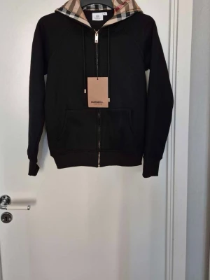 Svart hoodie från Burberry med dragkedja - Snygg svart hoodie från Burberry med dragkedja framtill och klassiskt rutigt foder i huvan. Hoodien har två fickor fram och ribbade muddar vid ärmar och nederkant. Perfekt för en stilren och avslappnad look. Materialet känns mjukt och bekvämt.