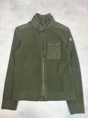 Grön fleecejacka från Moncler - Mörkgrön fleecejacka från Moncler med hög krage, bröstficka med knapp och dragkedja framtill. Jackan har en diskret Moncler-logga på ärmen och detaljer i nylon på axlar och ficka. Perfekt för lager-på-lager under kyliga dagar.