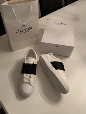 Valentino Garavani Open sneakers vit/svart - Säljer ett par Valentino Garavani Open sneakers i vitt skinn med bred svart rem över vristen. Klassisk rund tå, vita snören och cool räfflad sula. Kommer med originalkartong, dustbag och tags. Perfekta för dig som vill ha en clean men lyxig look.