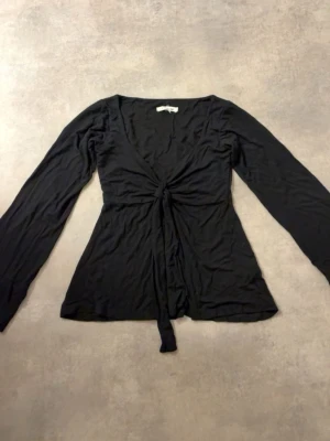 Svart omlottopp med knyt - Svart omlottopp med långa ärmar och knyt framtill. Toppen har en djup v-ringning och är tillverkad i ett mjukt, stretchigt material som ger en smickrande passform. Perfekt att styla med jeans eller kjol.