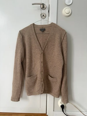 Beige kofta från Acne Studios - Snygg beige kofta från Acne Studios med v-ringning och knäppning framtill. Koftan har två fickor fram och är tillverkad i mjukt material som känns lyxigt. Perfekt att slänga över en t-shirt eller skjorta för en clean look.
