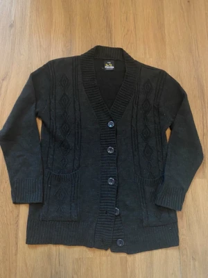 Svart Cardigan - Mysig svart stickad kofta från Florida med ribbade muddar, stora knappar och två framfickor. Koftan har ett diskret mönster längs framstyckena och djup V-ringning. Perfekt att slänga på sig när det är kyligt ute.