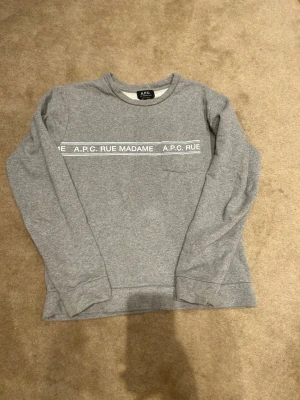 Grå sweatshirt från A.P.C. - Snygg grå sweatshirt från A.P.C. med trycket 'A.P.C. RUE MADAME' över bröstet och en liten bröstficka. Tröjan har rund halsringning och långa ärmar. Pris kan diskuteras!! Vid minsta fundering är det bara att höra av sig!