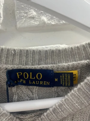 Grå kabelstickad tröja Polo Ralph Lauren - Snygg grå kabelstickad tröja från Polo Ralph Lauren. Tröjan har rund halsringning, långa ärmar och ribbade muddar vid ärmslut och nederkant. Perfekt för lager på lager och enkel att matcha med jeans eller kjol.