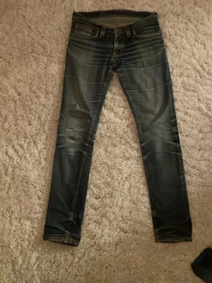 Nudie jeans - Säljer ett par slim fit blå jeans med slitningar och tvättade detaljer. Jeansen har klassisk femficksmodell och dragkedja. Snygga slitningar på ena benet ger en cool och avslappnad look. Perfekta till sneakers och hoodie.