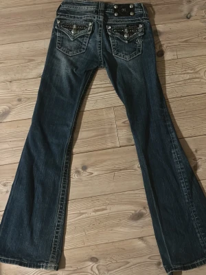 Mörkblå bootcut jeans från Miss Me - Säljer ett par mörkblå bootcut jeans från Miss Me med snygga detaljer på bakfickorna, inklusive nitar, broderier och glittriga stenar. Jeansen har klassisk femficksdesign och är gjorda i denim med slitningar för en cool look. Nypris: 2000 kr (Pm för intresse av mått)