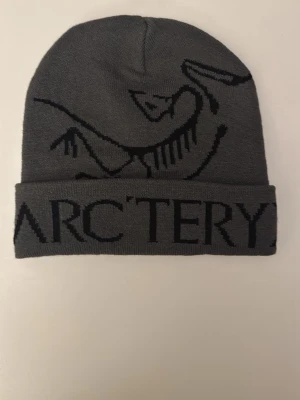 Grå mössa från Arc'teryx - Snygg grå mössa från Arc'teryx med svart logotyp och grafiskt mönster. Mössan är stickad och har uppvikt kant med tydlig text. Perfekt för dig som gillar stilrena accessoarer med outdoor-vibe.