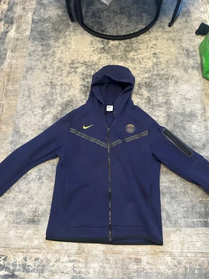 Blå Nike PSG hoodie med dragkedja - Snygg mörkblå hoodie från Nike x PSG med hel dragkedja, huva och fickor. Har PSG-logga på bröstet och gul Nike-swoosh. Detaljer i svart på bröstet och en extra ficka med dragkedja på ärmen. Perfekt för dig som gillar sportig stil.