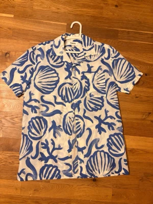 Vit och blå skjorta från Kappahl - Kortärmad skjorta från Kappahl i vit och blå färg med stort, grafiskt mönster av snäckor och havsväxter. Skjortan har klassisk krage, bröstficka och knappar framtill. Tillverkad i ett lätt och luftigt linnematerial, perfekt för varma dagar. Pris kan diskuteras!