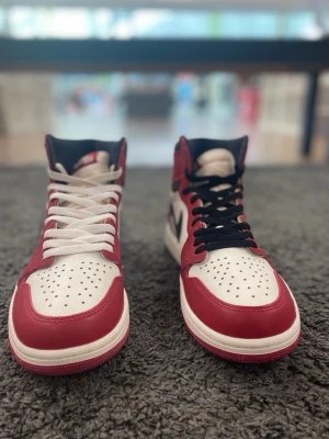 Air Jordan 1 High Lost and found Chicago  - Säljer ett par klassiska Air Jordan 1 High Chicago sneakers i rött, vitt och svart läder. Skorna har perforerad tå, svart swoosh på sidan och olika färg på skosnörena – ena vit och andra svart. Ikonisk high-top siluett med retrovibbar och tydliga Jordan-detaljer.