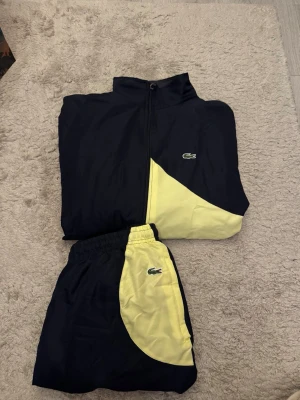 Lacoste träningsset blå/gul - Snyggt träningsset från Lacoste i mörkblått och ljusgult. Setet består av en zip-jacka och matchande byxor, båda med Lacoste-loggan broderad på bröstet och benet. 
