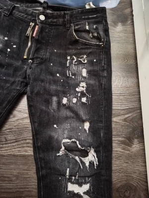 Svarta distressed jeans från Dsquared2 - Säljer ett par svarta jeans från Dsquared2 med slitningar, hål och färgstänk för en riktigt edgy look. Jeansen har dragkedja framtill, fem fickor och tydliga detaljer som förstärkta sömmar och Dsquared2-logga vid gylfen. Perfekta för dig som gillar streetwear och statement-plagg .Helg Rea nu 👍