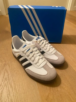 Adidas Samba OG vita och svarta sneakers - Klassiska Adidas Samba OG sneakers i vitt läder med grå mockadetaljer och svarta ränder på sidorna. Låg modell med gummisula och blå logga på plösen. Perfekta för en stilren och sportig look.