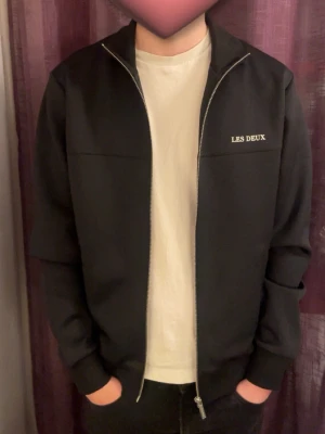 Svart tröja från Les Deux med dragkedja - Stilren svart tröja från Les Deux med silverfärgad dragkedja och diskret logga på bröstet. Jackan har en klassisk passform, ribbade muddar och är perfekt för dig som gillar en clean look. Passar till många olika outfits. Nypris 1300kr. Stl M men passar även S. Finns även tillhörande byxor. 