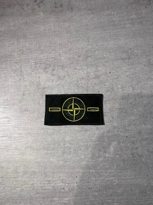 Stone Island svart tygmärke - Säljer ett svart tygmärke från Stone Island med det klassiska kompassloggan broderad i gult och grönt. Märket är rektangulärt och har detaljerade broderier, perfekt att sy eller fästa på jackor, tröjor eller väskor.