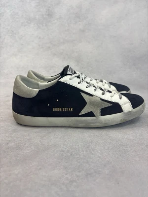 Svarta Golden Goose sneakers med stjärna - Snygga svarta och vita sneakers från Golden Goose med ikonisk silverfärgad stjärna på sidan. Skorna har vita snören med text, vit sula och detaljer i mocka och läder. GGDB/SSTAR tryckt i guld på sidan. Perfekta för en trendig streetstyle-look.