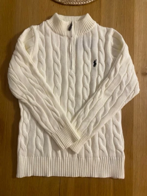 Vit kabelstickad tröja Polo Ralph Lauren - Kabelstickad vit tröja från Polo Ralph Lauren med halv dragkedja och ribbad hög krage. Tröjan har det klassiska mörkblå broderade logotypen på bröstet och ribbade muddar vid ärmslut och nederkant. Perfekt för en stilren och tidlös look.