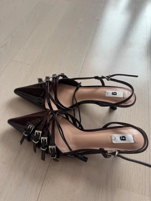 Vinröda lackpumps med spännen - Säljer ett par vinröda pumps i lack med spetsig tå och flera tunna remmar med silvriga spännen. Skorna har smal klack och justerbara ankelband för extra detalj. Perfekta för dig som gillar edgy och trendig stil.