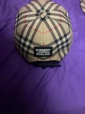 Burberry beige rutig keps - Säljer en klassisk Burberry keps i beige med det ikoniska rutmönstret i svart, vitt och rött. Kepsen har en böjd skärm och justerbar rem bak. Märkt med 'BURBERRY LONDON ENGLAND' på baksidan. Perfekt accessoar för en trendig streetwear-look.