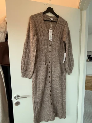 Stickad lång klänning/ kofta happy Holly  - Lång, stickad kofta i beige melange från Happy Holly. Koftan har v-ringning, knappar hela vägen framtill, två stora fickor och ribbade muddar vid ärmslut. Materialet är mjukt och fluffigt, perfekt för lager på lager och en chill look. Storlek 36/38 och köptes för 699kr, prislappar sitter kvar