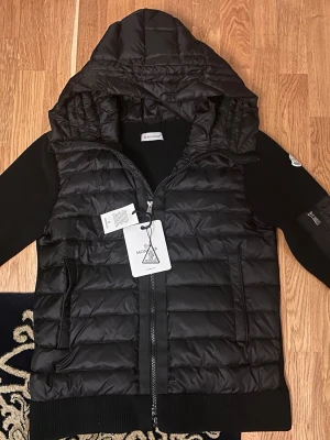 Moncler cardiganjacka i storlek S - Helt Ny - Denna Moncler cardiganjacka i storlek S kombinerar funktion och stil. Den har en quiltad design med en huva för extra värme och komfort. Perfekt för kalla dagar, och med sin svarta färg passar den både till vardags och mer avslappnade tillfällen. Jackan har också en praktisk armficka och en trendig siluett som gör den till ett utmärkt val för både stilmedvetna och praktiska användare.