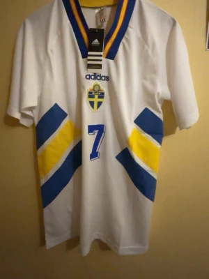 Vintage Sverige-tröja - Larsson #7 - klassisk Sverige-tröja från Adidas i storlek M med Larsson #7 på ryggen. Den här tröjan är en riktig retro-pärla och perfekt för alla fotbollsälskare och Larsson-fans. Den är i bra skick och redo att bäras eller samlas på. Tröjan har en snygg design med Sveriges färger och är ett måste för alla fans.