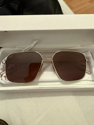 CHIMI solglasögon  - Snygga solglasögon från CHIMI med fyrkantiga bruna glas och tunn guldfärgad metallbåge. Trendig design som ger en clean och modern look. Perfekta för dig som vill sticka ut med stilrena accessoarer.