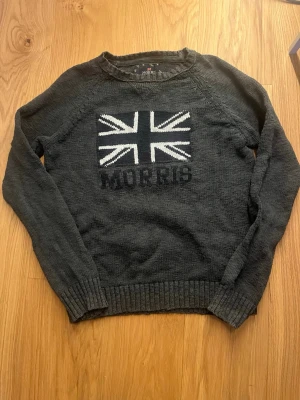 Mörkgrå stickad tröja från Morris - Snygg mörkgrå stickad tröja från Morris med Union Jack-flagga och logotyp framtill. Tröjan har rund halsringning, långa ärmar och ribbstickade muddar vid ärmslut och nederkant. Perfekt för dig som gillar klassisk och stilren design.