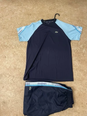 Blått Lacoste träningsset - Snyggt träningsset från Lacoste med t-shirt och shorts i mörkblått och ljusblått. T-shirten har raglanärmar och logga på bröstet, medan shortsen har elastisk midja och matchande mönstrad detalj. Perfekt för sport eller chill.