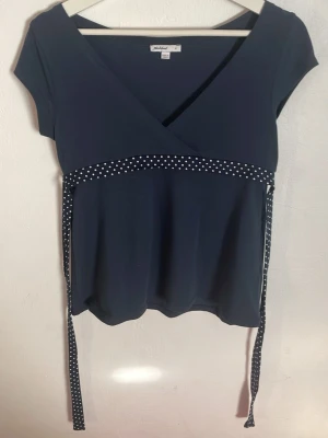 Mörkblå omlottopp med prickigt band - Mörkblå omlottopp med korta ärmar och ett dekorativt midjeband i mörkblått med vita prickar. Toppen har v-ringning och knytband i midjan för en snygg siluett. Perfekt för dig som gillar klassiska detaljer och enkel stil.