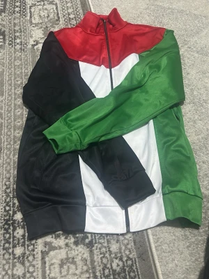 Färgglad träningsjacka med dragkedja - Snygg träningsjacka i glansigt material med färgblock i rött, grönt, svart och vitt. Jackan har hög krage, hel dragkedja framtill och muddar vid ärmslut och nederkant. Perfekt för dig som vill sticka ut på gymmet eller ute.