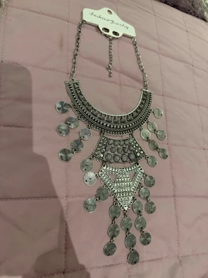 Statementhalsband i silverfärg - Unikt statementhalsband i silverfärgad metall med stora geometriska former och hängande runda myntliknande detaljer. Halsbandet har en bohemisk och trendig känsla, perfekt för att lyfta vilken outfit som helst. Justerbar kedja för bästa passform.