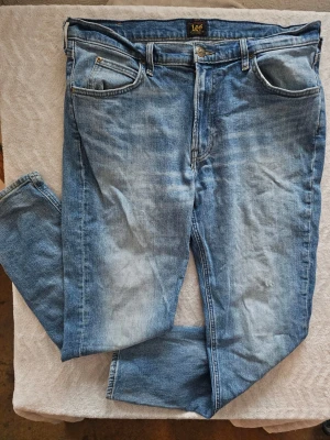Blå raka jeans från Lee - Säljer ett par klassiska blå jeans från Lee med raka ben och fem fickor. Jeansen har en snygg tvättad look och Lee-logga på knapp och etikett. Tillverkade i slitstarkt denimtyg, perfekta för en avslappnad stil.