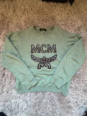Turkos Grön sweatshirt från MCM Storlek S - Cool grön sweatshirt från MCM med stort broderat logotyptryck framtill. Tröjan har rund halsringning och långa ärmar, perfekt för en avslappnad och trendig look. Inte för tjock och väldigt skön, perfekt för kommande säsongerna. Bra skick.
