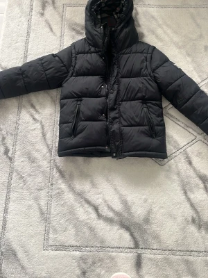 Svart pufferjacka från Burberry - Snygg svart pufferjacka från Burberry med huva och dragkedja framtill. Jackan har två snedställda fickor med dragkedja och quiltad design. Perfekt för kalla vinterdagar och ger en trendig look. Materialet är slitstarkt och håller värmen bra.