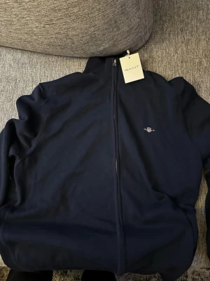 Mörkblå ziptröja från GANT - Snygg mörkblå ziptröja från GANT med hög krage och dragkedja framtill. Tröjan har ett diskret GANT-logo broderat på bröstet och ribbade muddar vid halsen. Perfekt för en clean och stilren look.