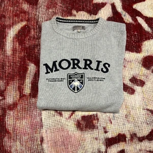 Grå stickad tröja från Morris - Snygg grå stickad tröja från Morris med svart broderad logga och emblem på bröstet. Tröjan har rund halsringning med ribbade muddar och nederkant. Perfekt för dig som gillar klassisk och stilren design.