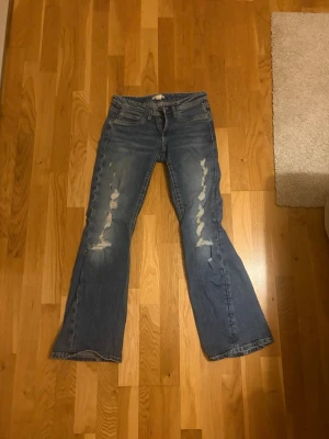Blå bootcut jeans med slitningar - Säljer ett par blå bootcut jeans från Gina tricot med tydliga slitningar framtill och klassiska fem fickor. Jeansen har en avslappnad passform och är tillverkade i denim. Perfekta för en cool och trendig look.