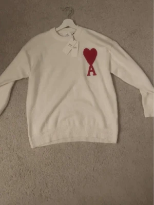Vit stickad tröja från Ami - Säljer en vit stickad tröja från Ami pari med rund halsringning och ribbade muddar. Tröjan har ett rött hjärta och bokstaven 'A' broderat på bröstet. Perfekt för dig som gillar stilrena plagg med en twist.