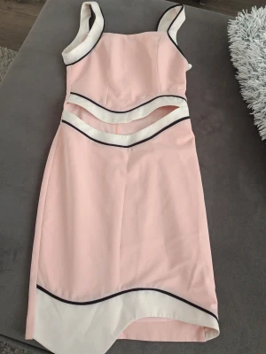Ljusrosa klänning med cut out-detaljer - Säljer en oanvänd ljusrosa klänning med vita och svarta kontrastkanter. Klänningen har breda axelband, cut out-detaljer i midjan och asymmetrisk nederkant. Materialet känns som polyester och modellen är figurnära med en modern twist.