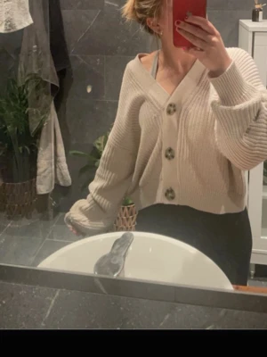 Beige kofta - Kort, beige kofta med ribbad struktur och djup V-ringning. Tre stora, marmorerade knappar framtill. Perfekt att slänga över en topp för en avslappnad men snygg look.